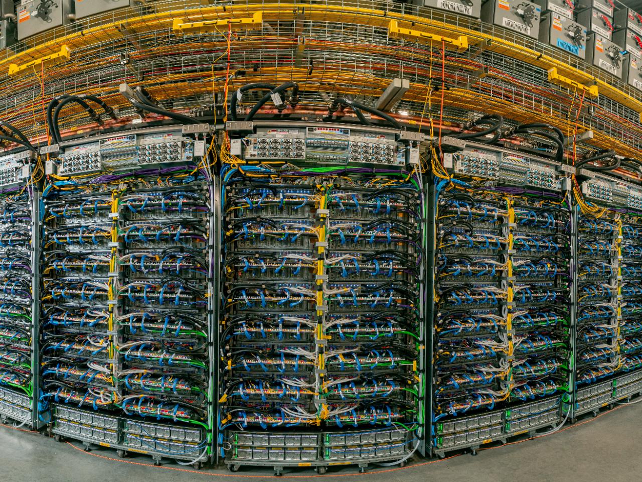 Wide-angle photo of a curved wall of Ironwood TPU racks packed with blue and yellow cables, under yellow overhead cable trays in a Google data center.