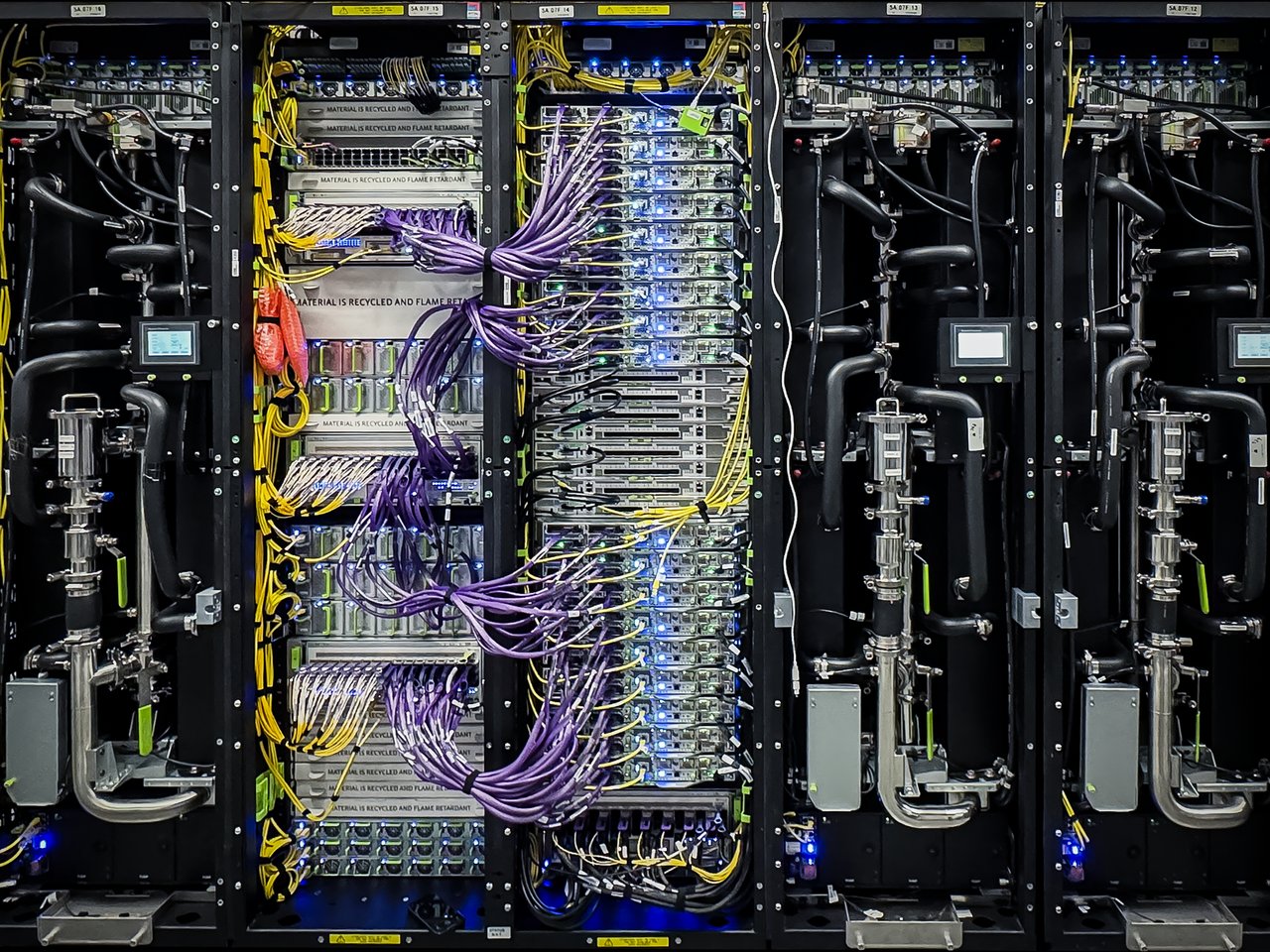Photo of an MTIA 400 rack with its doors open: center racks of server trays wired with purple and yellow cables, between black liquid cooling racks with silver pipes.